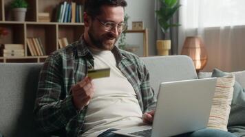 Happy smiling client buyer Caucasian man male guy at home with credit card and laptop computer shopping buying online purchase check money financial account order food delivery payment internet bank video