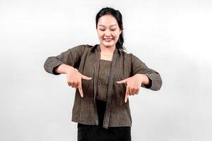 An Indonesian woman wearing traditional lurik attire smiles brightly while pointing downwards with both hands, showcasing cultural pride and enthusiasm against a neutral background. photo