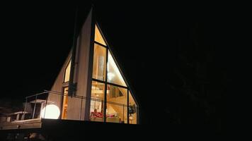 Night view of modern A-frame cabin with large illuminated windows, cozy indoor holiday setting. video