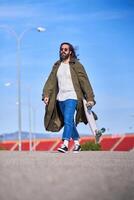 Trendy hipster man in raincoat walking with skateboard photo