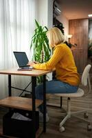 Young woman working at a cozy desk in a warm-toned room surrounded by greenery while typing on a laptop during the afternoon photo