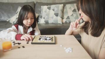 Asian family, mum, and kid fun playing Go, checkers board game on table in domestic room together with love and cheerful, happy weekend relaxing hobby of child and parent, concentrate and compete. photo