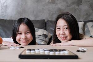 Portrait of Asian mum, and kid fun playing Go, checkers board game on table in home living room with love and cheerful, happy weekend family times relaxing hobby, smile and look at camera together. photo