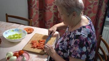 An elderly woman is preparing food in the kitchen. She is cutting a tomato salad of vegetables and fruits. video