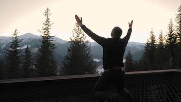 Man kneeling with raised hands in prayer before majestic mountains at sunset, embracing the divine presence in nature, snowy peaks, evergreen trees, and a peaceful landscape of faith and gratitude video