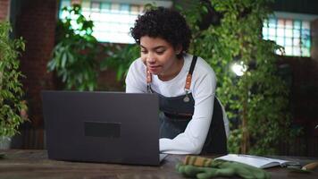 African woman florist gardener working in botanical store using laptop order plants communicating on Internet with customer. Female small business owner working at flower shop. Running of own business video