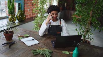 African woman florist gardener working in botanical store using laptop order plants by phone communicating with customer. Female small business owner working at flower shop. Running of own business video