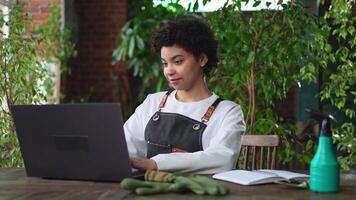 African woman florist gardener working in botanical store using laptop order plants communicating on Internet with customer. Female small business owner working at flower shop. Running of own business video