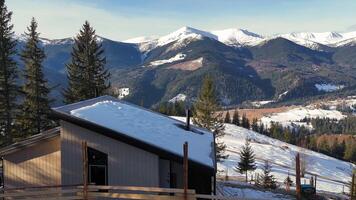 Cozy wooden cabin with a snow-covered roof in the mountains, surrounded by evergreen trees and video