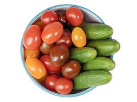 Small colorful cherry tomatoes and fresh green cucumbers overflowing in bowl with blue border, top view, abundant harvest concept, isolated on white background, photo