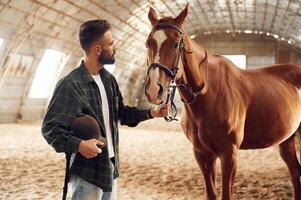 Standing and holding animal. Young man with a horse is in the hangar photo
