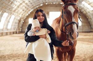 Mother with, baby on the hands is standing indoors with horse in hangar photo
