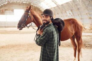 Holding the ,saddle in hands.. Young man with a horse is in the hangar photo