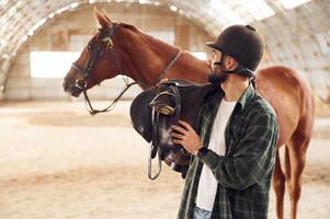 Holding the saddle in hands. Young man with ,a horse is in the hangar photo