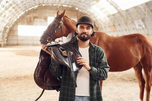 With saddle in hands. Young man with a horse ,is in the hangar photo
