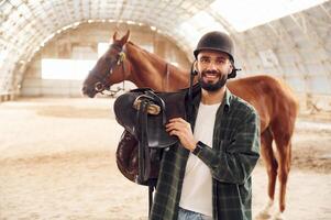 With saddle in hands. Young man with ,a horse is in the hangar photo