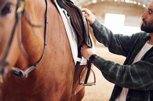 Process of preparation, the animal for a ride. Young man with a horse is in the hangar photo