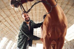 Smiling ,and taking care of animal. Young ,man with a horse is in the hangar photo