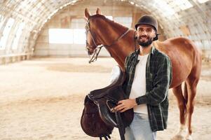 Holding ,the saddle in hands. Young ,man with a horse is in the hangar photo