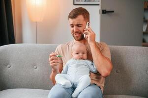 Calling doctor. With thermometer.. Father with his newborn baby is indoors. Conception of single dad photo