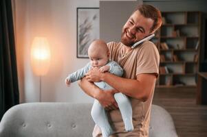 Holding kid and talking by phone. Father with his newborn baby is indoors. Conception of single dad photo