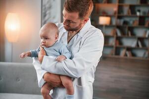 Looking ,at the kid. Doctor standing and holding newborn baby in hands indoors at apartment photo