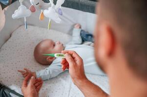 Holding, thermometer, kid in the cradle. Father with his newborn baby is indoors. Conception of single dad photo