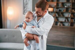 Looking at the kid. Doctor standing and holding ,newborn baby in hands indoors at apartment photo