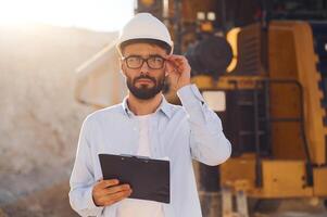 In hard hat, eyewear, and with notepad. Man in uniform is working in the quarry at daytime photo