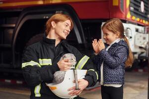 Cat is sitting, in the helmet, kid is happy, clapping her hands. Firefighter woman in uniform is with a little girl photo