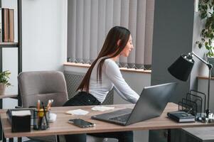 Young beautiful woman in white clothes is in the office and sitting by the table with laptop photo