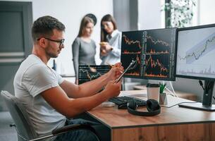 Stock, broker is sitting by computer and working with exchange technology. Women is standing and talking behind photo