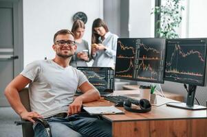 Stock broker, is sitting by computer and working with exchange technology. Women is standing and talking behind photo