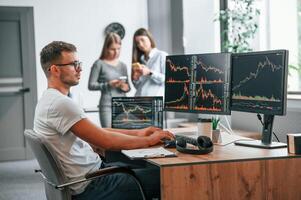 Stock broker ,is sitting by computer and working with exchange technology. Women is standing and talking behind photo