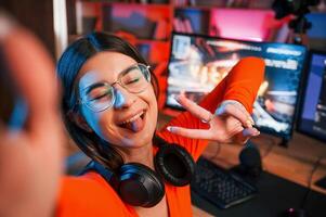 Smiling, making victory ,gesture and doing selfie. Young beautiful game streamer is indoors with pc photo