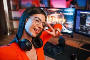Smiling,, making victory gesture and doing selfie. Young beautiful game streamer is indoors with pc photo