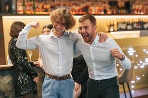 Two men are standing together in formal ,clothes on the party event. Friends are behind photo