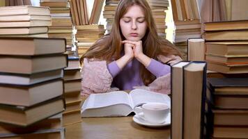 A girl reads a book in the library. Stacks of books and cup of tea on table. The girl is tired of studying. Concept of Reading and Education. Ultra 4K video