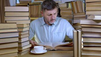 A bearded man reads a book in the library. Stacks of books and cup of tea on table. Concept of Reading and Education. video