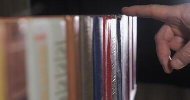 A mans hand is looking for the right book among a collection of volumes of books on a wooden bookshelf. Close Up. Shallow depth of field. Ultra 4K video
