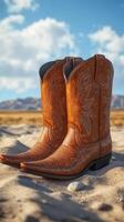 Brown cowboy boots rest on sandy terrain with mountains in the background under a blue sky photo