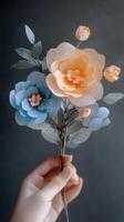 Handcrafted paper flowers in soft blue and peach tones with delicate leaves held against a dark background photo