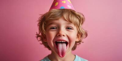 Child celebrates birthday with party hat, playful expression, and sticky tongue against a pink background photo