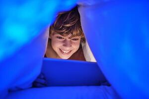 Joyful boy browsing tablet under blanket in late evening photo