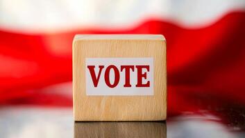 Vote block on table with flag background. A wooden block marked with the word vote rests on a reflective surface in front of a red and white flag backdrop. photo