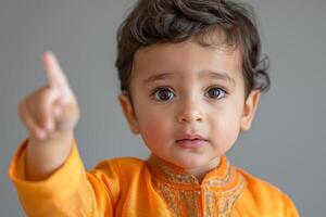 A young boy of South Asian descent, wearing an orange traditional outfit, curiously gestures with his finger while looking at the camera. photo