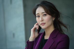 A thoughtful Asian woman in a stylish purple blazer speaks on her smartphone, embodying modern confidence and grace. photo