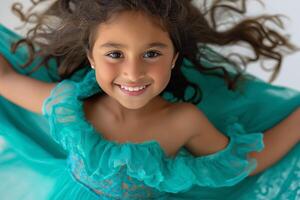 A joyful young girl of Middle-Eastern descent in a flowing turquoise dress, smiling brightly against a white background. photo