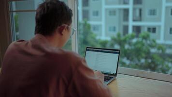 A young adult, seated at a desk in front of a window with a view of the city, diligently works on their laptop.Displays a web page with a text interface using a modern chat tool. video