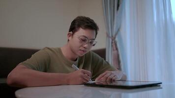 A young man wearing glasses takes notes intently while sitting at a desk, focused on his work. His determined expression conveys his concentration and dedication. video